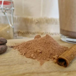 Poudre pour chocolat chaud accompagnée de fève de cacao et d'un baton de cannelle sur une planche en bois.
